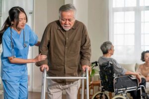 Three older adults smiling together in a cozy living room, with a caregiver nearby offering gentle assistance.