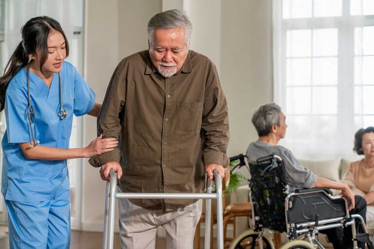 Three older adults smiling together in a cozy living room, with a caregiver nearby offering gentle assistance.